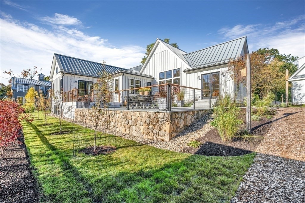 A white farmhouse with a raised terrace held by a fieldstone wall, cedar handrails with cable infill, and new lawn and planting in front.