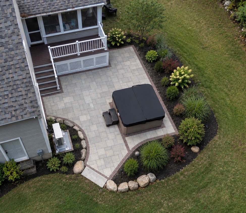 Overhead view of a paver patio with a hot tub, wrapped by planting beds and a boulder border beside the house.