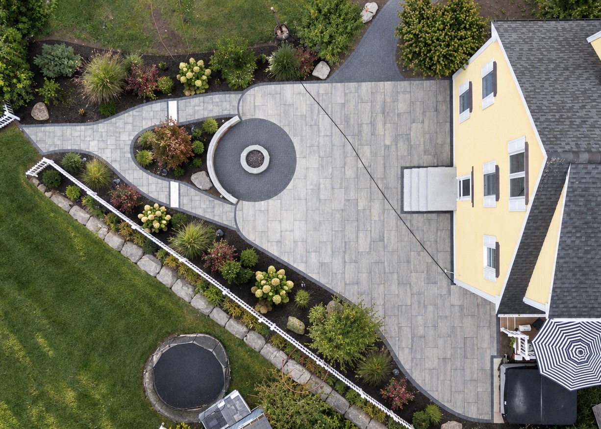 Overhead view of a paver patio with a circular fire pit and a curved seating wall, and a walkway looping through planted beds.