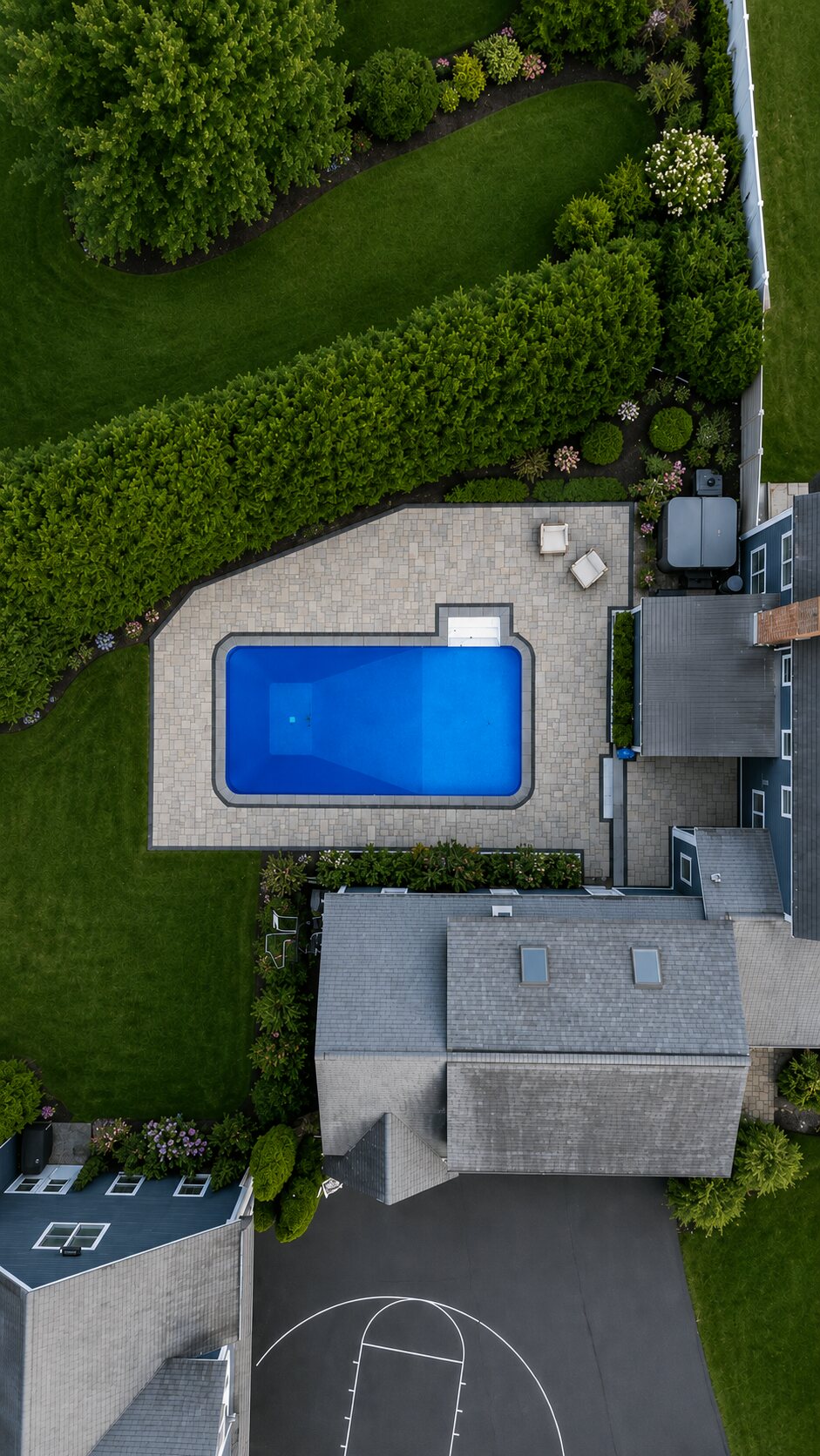 Overhead view of a rectangular in-ground pool set in a paver deck, with a hedge, planting beds and lawn around it.